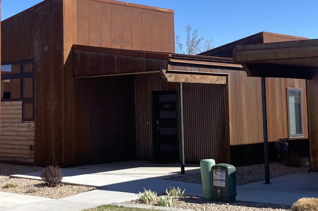 A modern house with a rusty, weathered metal exterior, flat roof, and geometric design. The entrance is shaded by an overhang, and small desert plants line the concrete walkway leading to the front door.