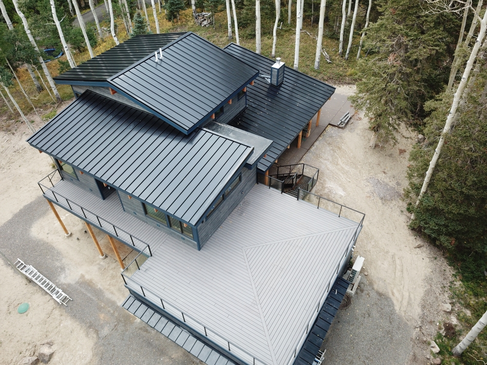 Aerial view of a modern house with a flat, dark roof and tall windows, nestled among tall, slender trees. The structure features a spacious deck and a surrounding gravel area.