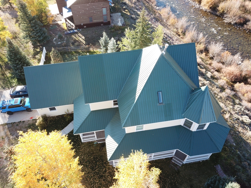 Aerial view of a house with green metal roofing and beige siding, situated near a river and surrounded by autumn trees. A blue vehicle is parked in the driveway, and another building is visible in the background.