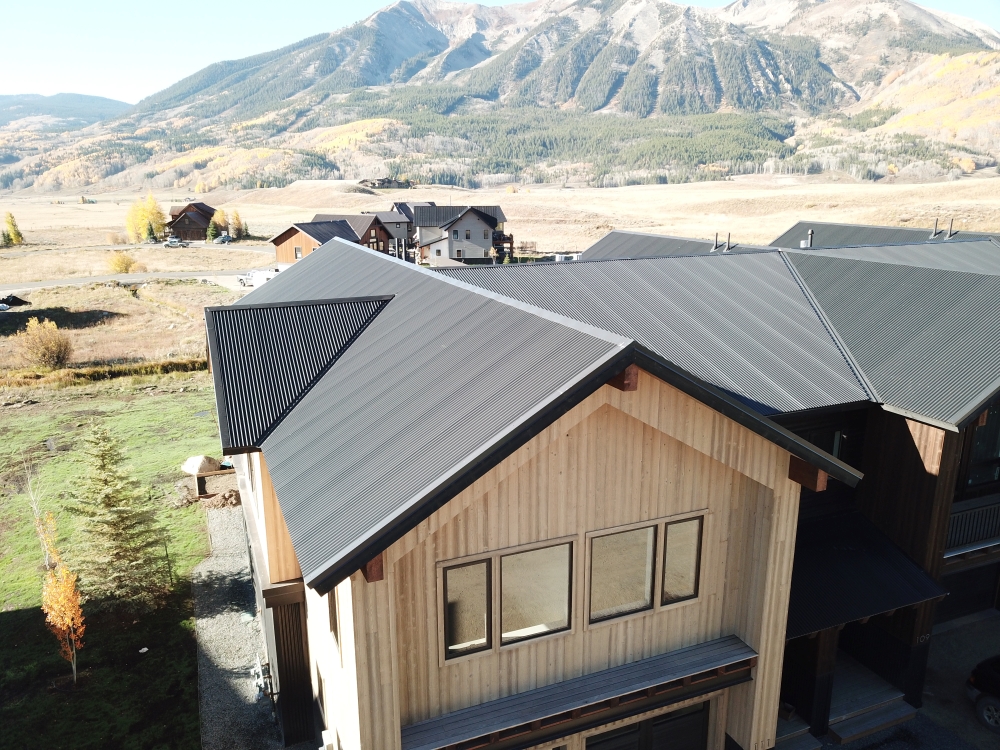 Aerial view of a modern, two-story house with dark metal roof and light wood siding. The house is set against a mountainous landscape, with other houses nearby and autumn trees scattered throughout the scene.