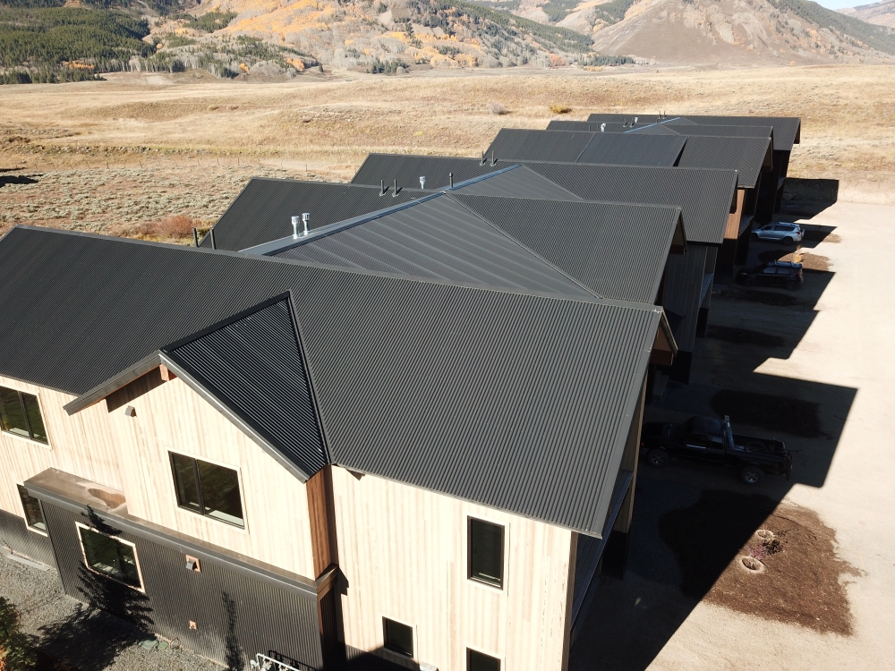 Aerial view of a row of modern houses with dark metal roofs and light wooden walls in a rural landscape. The background shows mountains and a mix of grassy and wooded terrain under a clear sky.