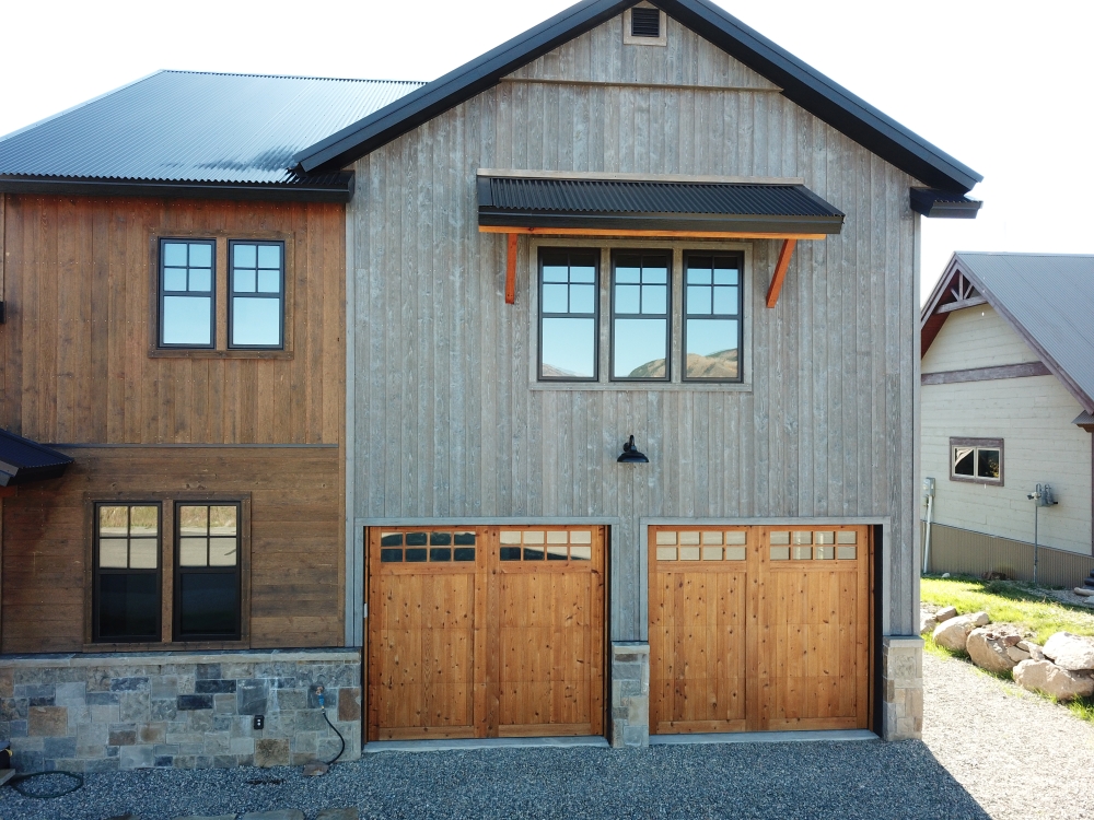 A rustic house with a two-car wooden garage. The building features a mix of wood and stone materials with large windows. The roof is metal, and the driveway is gravel. The overall style is modern farmhouse.