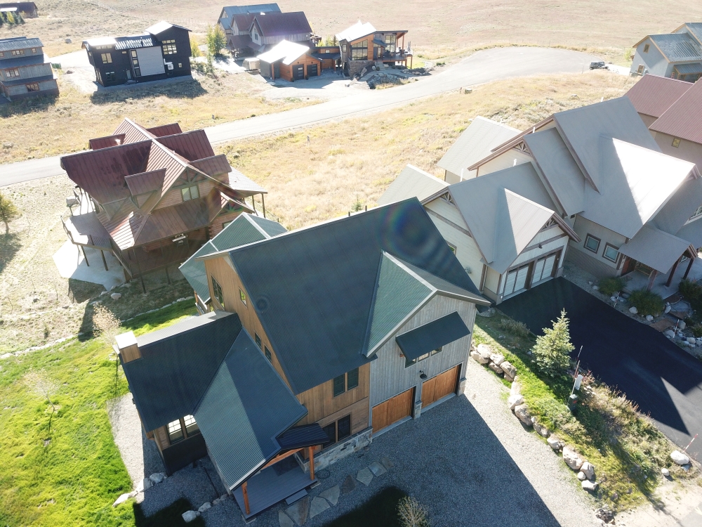 Aerial view of a residential neighborhood with modern houses. The foreground features a large, multi-story house with a dark roof and wooden accents. Other houses and a road are visible in the background, surrounded by dry grass and shrubs.