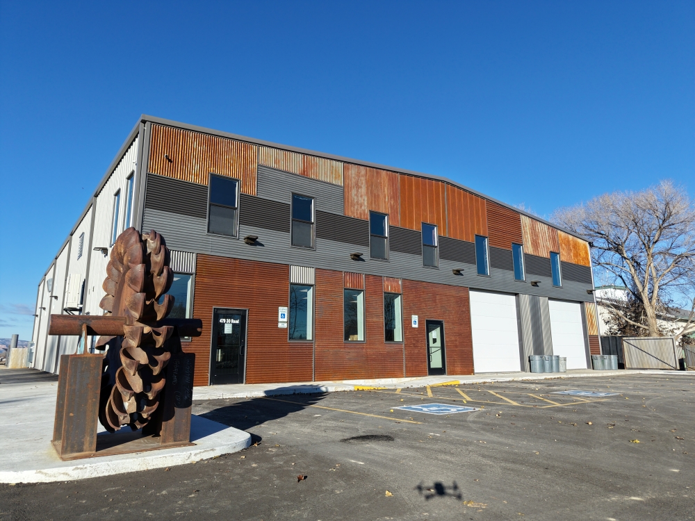Warehouse-style building with a mix of metal and wood panels on blue sky. Includes parking spaces and a sculpture of a large pine cone on a stand near the entrance. The building has ground-level entrances and windows.