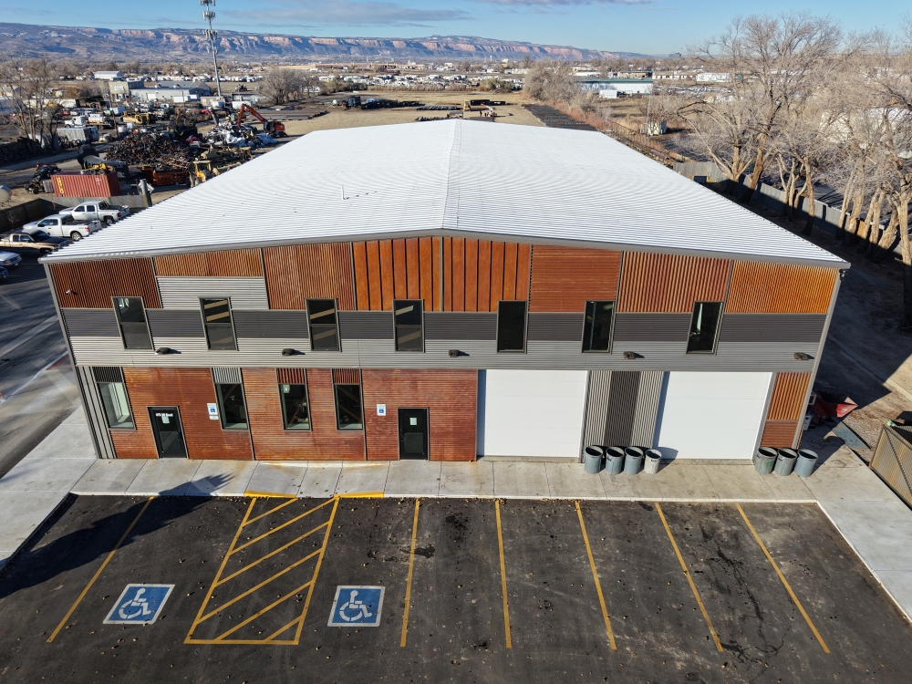 Aerial view of a large rectangular industrial building with corrugated metal siding in various shades of brown and gray. The lot features two handicap parking spaces in front and surrounding industrial areas in the background.