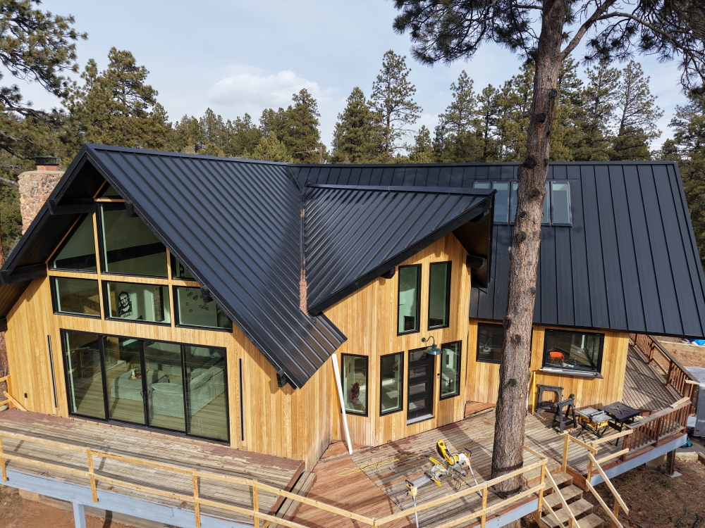A modern wooden cabin with a steep, dark metal roof is surrounded by trees. Large windows line the front, and there's a wrap-around deck with construction tools. Tall pine trees are visible in the background under a partly cloudy sky.