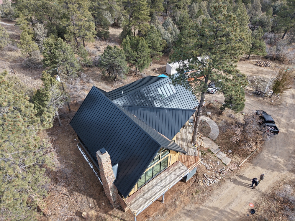 Aerial view of a modern cabin with a dark metal roof and large windows, surrounded by pine trees. A dirt road leads to the house, with two people walking nearby. The landscape is a mix of dry earth and scattered greenery.