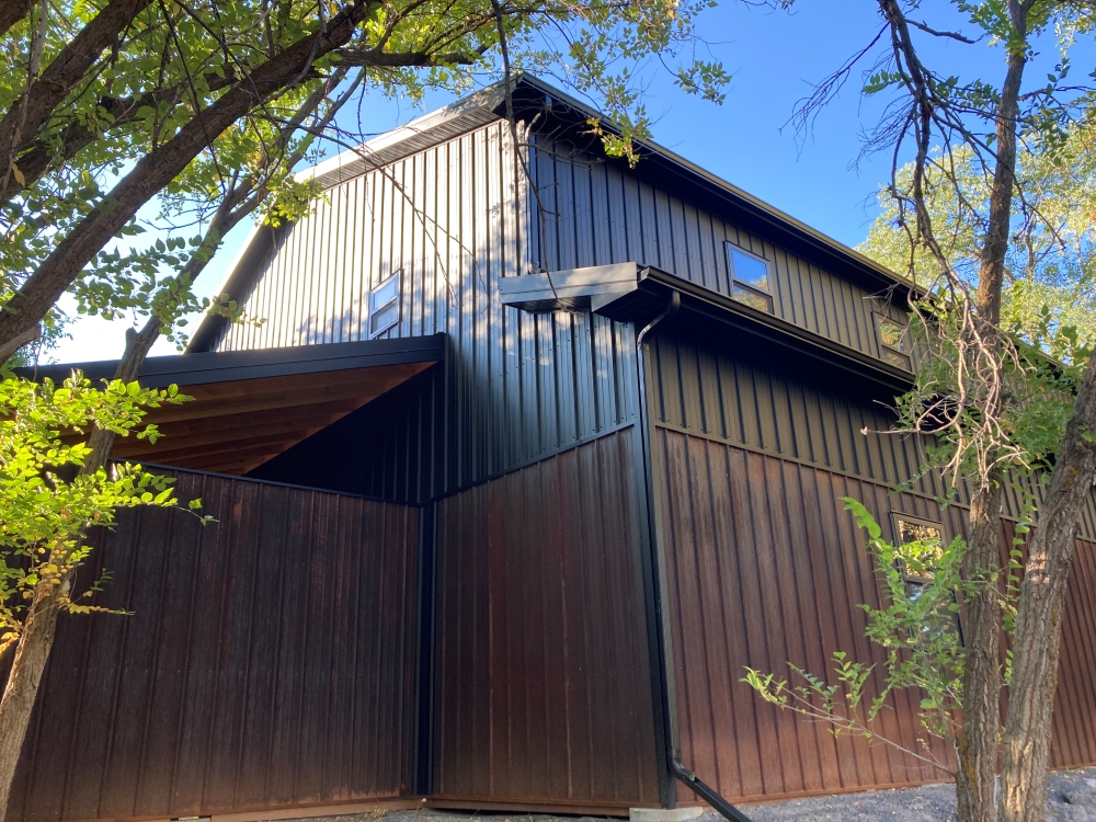 Modern two-story house with dark wood and metal siding, surrounded by leafy trees. The structure features sharp angles and a sloped roof, under a clear blue sky.