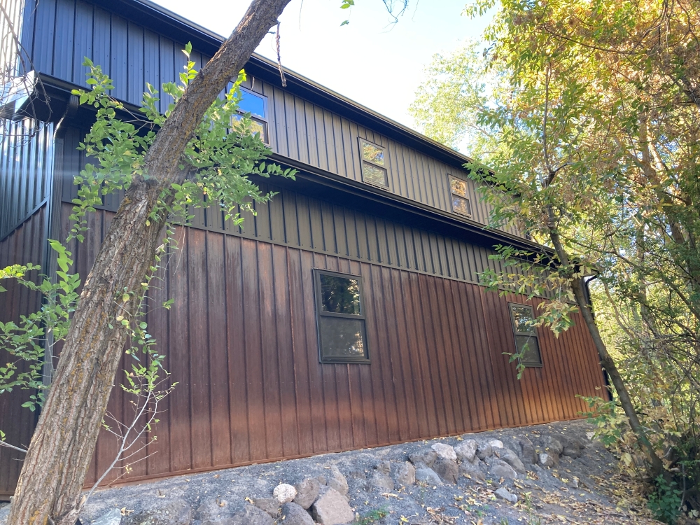 Two-story building with dark brown and black siding, nestled among trees. The structure has several windows visible on both floors and is partially surrounded by rocks and foliage. Sunlight filters through the leaves, casting dappled shadows.