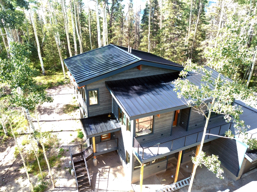 Aerial view of a modern, dark-colored house with a metal roof, surrounded by tall trees and a forest. The house has multiple levels, large windows, and a wooden staircase leading to an elevated entrance. Bright daylight illuminates the scene.