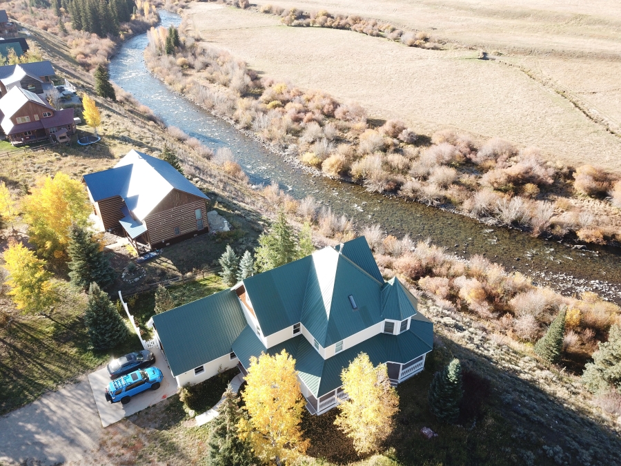 Aerial view of a house with a green roof and a parked blue vehicle beside it. Nearby, there's a river bordered by trees with autumn foliage and several other houses are visible. The landscape is mostly flat with open fields.