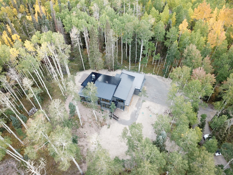Aerial view of a modern, dark-colored house with a metal roof surrounded by a dense forest. The trees have a mix of green and yellow leaves, indicating early autumn. A clear driveway leads to the house, and there is a small clearing around the building.