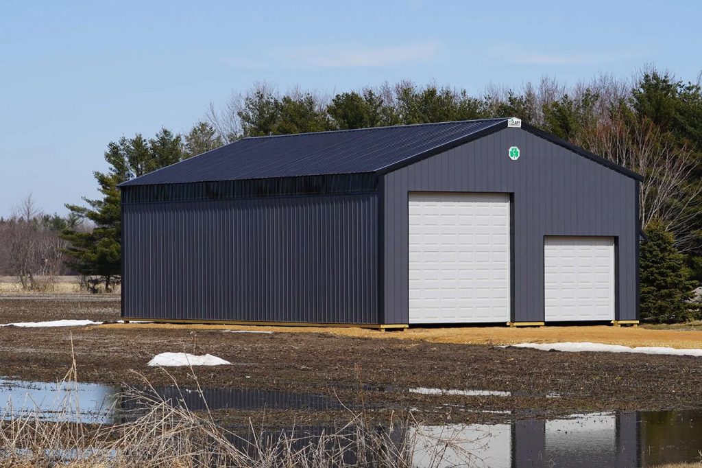 A gray metal storage building with a dark roof and two white roll-up doors stands on a dirt lot, surrounded by puddles, patches of melting snow, and trees in the background under a blue sky.