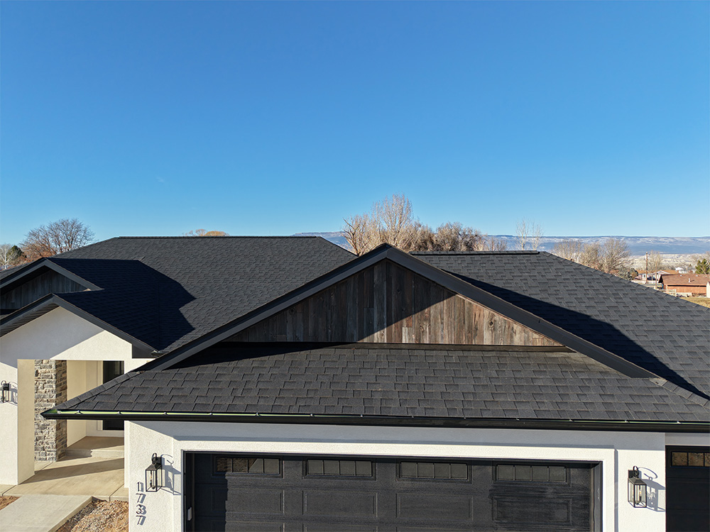 A modern house with dark shingle roofing and light-colored exterior walls, photographed on a clear day with a blue sky and some trees in the background.