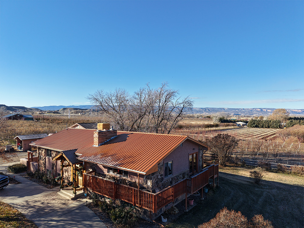 A single-story house with a red roof and large deck sits in a rural area, surrounded by fields, trees, and distant mountains under a clear blue sky.