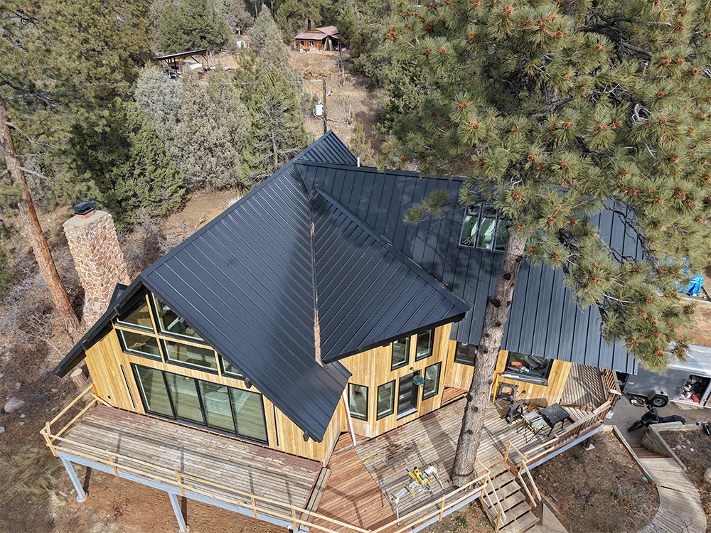 Aerial view of a modern wooden house with a dark metal roof, large windows, and a wraparound deck, surrounded by trees in a forested area.
