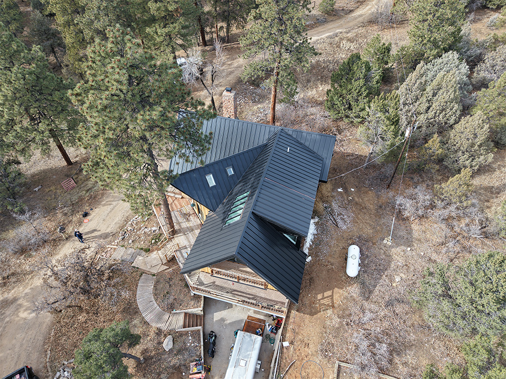 Aerial view of a house with a modern black metal roof surrounded by pine trees and dirt paths; vehicles and outdoor structures are visible near the house.