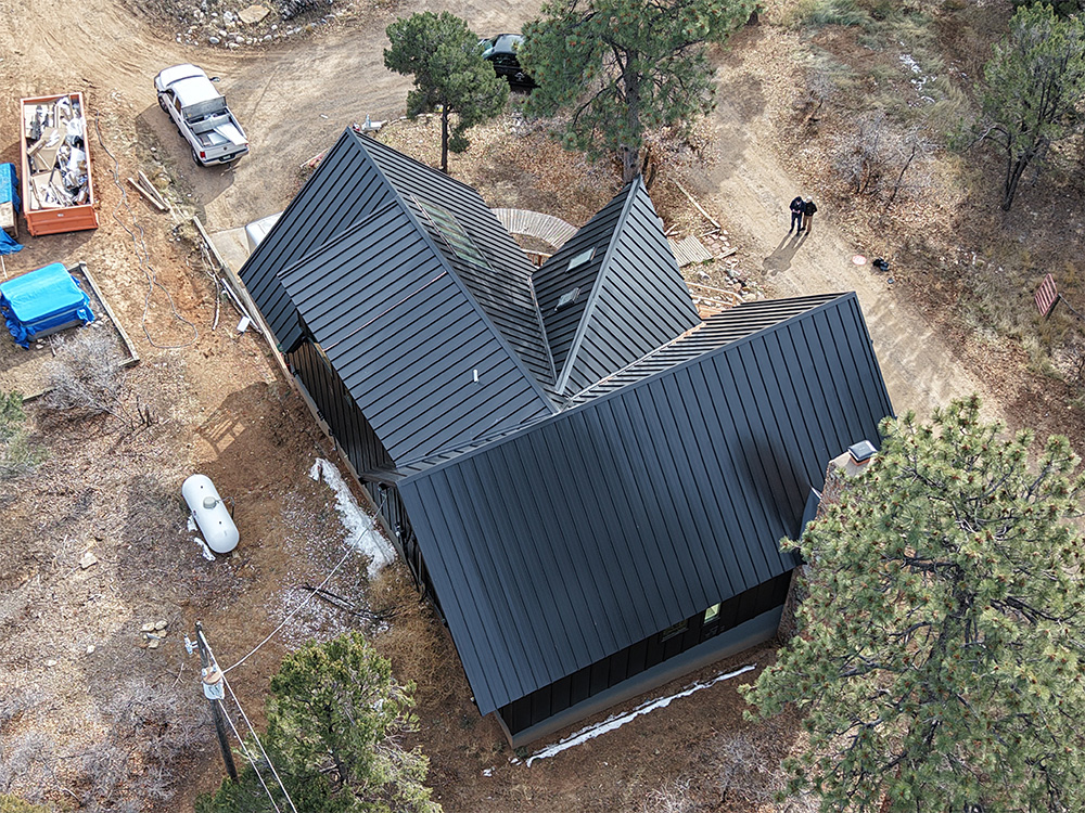 Aerial view of a modern house with a black metal roof surrounded by trees and dirt paths. Nearby are a parked car, a dumpster, scattered construction materials, and two people standing on the driveway.
