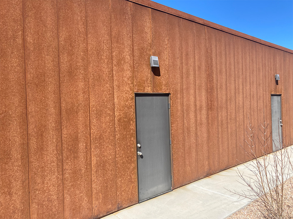 A modern building with a rust-colored metal exterior features two gray metal doors and wall-mounted lights, set against a clear blue sky. A bare shrub is visible near the right door on a paved walkway.