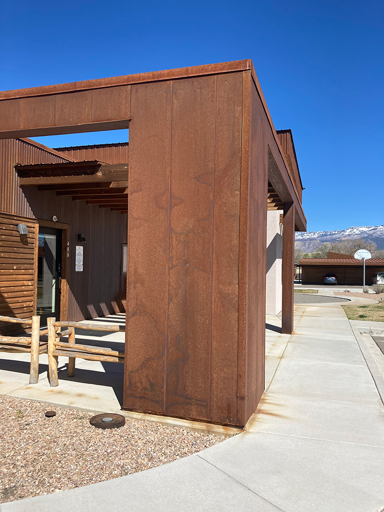 A modern building with rust-colored metal siding, wooden accents, and a sidewalk curving around it. There are mountains and a basketball hoop visible in the background under a clear blue sky.
