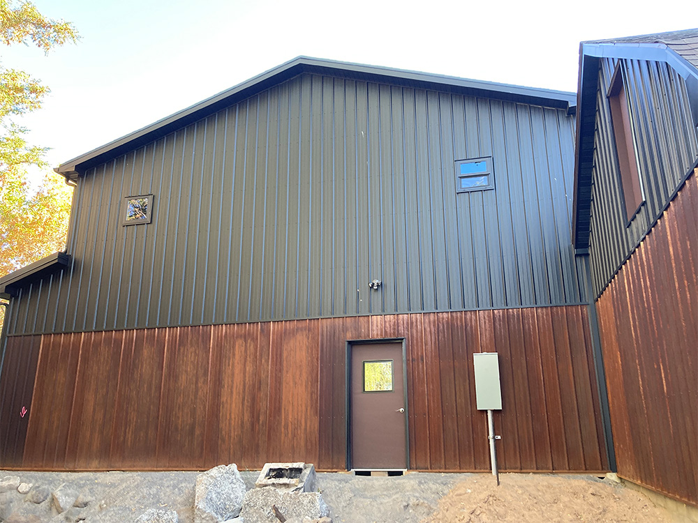 A metal-sided building with a sloped roof, two small windows, and a brown door set in a wooden lower facade. There are rocks and dirt in front, and an electrical box mounted on the wall.