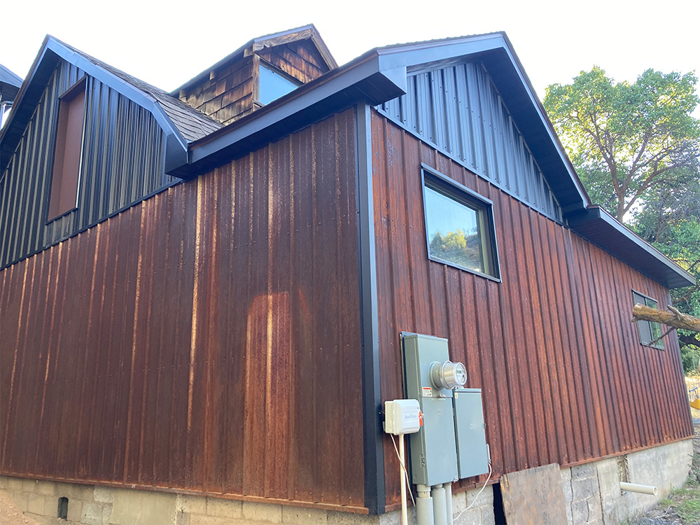 A house with a mix of rusted metal and dark siding, featuring rectangular windows, electrical meters, and surrounded by trees. The lower part of the building is concrete.