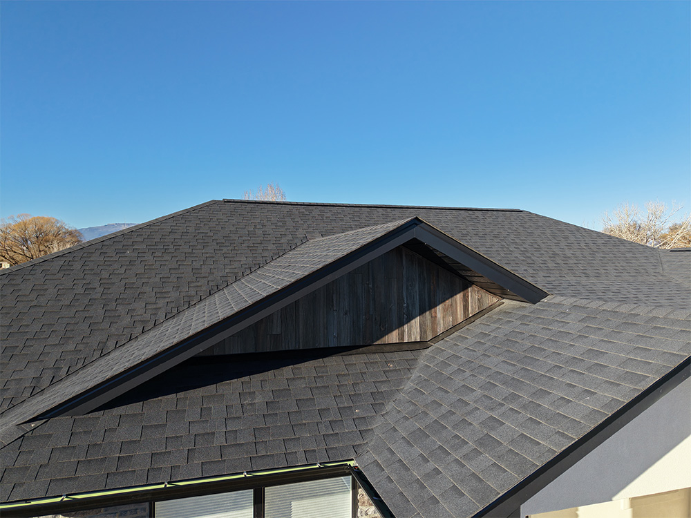 A modern house roof with dark gray asphalt shingles and sharp angles, set against a clear blue sky. Some tree branches are visible in the background.