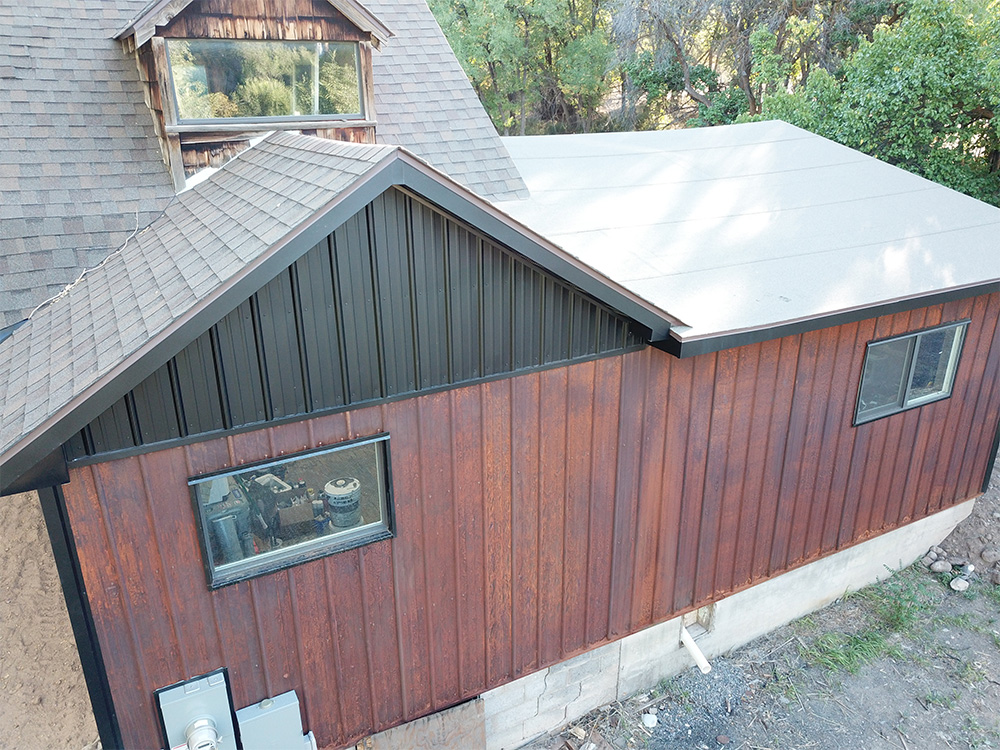 A wooden house with a brown shingle roof and an attached extension featuring vertical wooden siding, black trim, and large windows, surrounded by trees and some exposed ground.