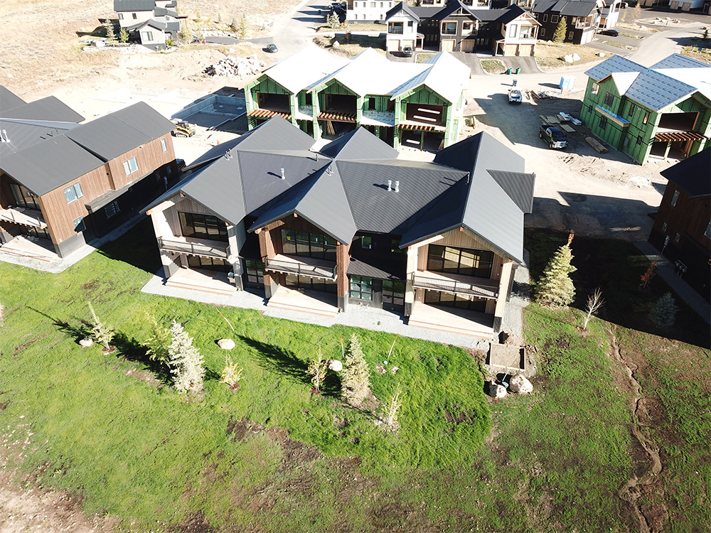 Aerial view of modern, dark-roofed duplex houses with balconies, surrounded by green lawn and small trees; other similar homes and construction area visible in the background.
