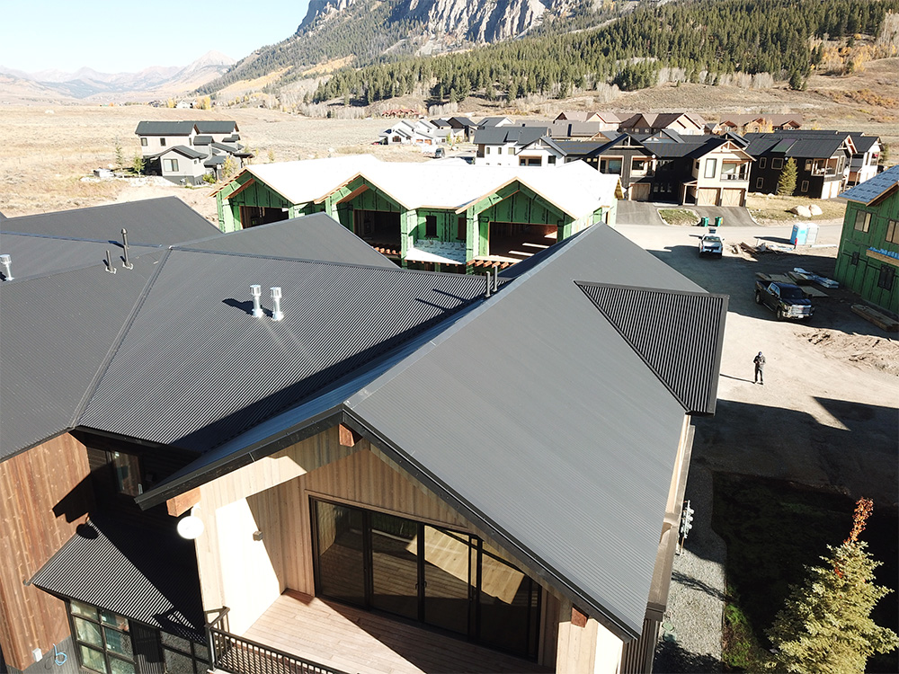 Aerial view of modern houses with dark metal roofs in a mountain neighborhood; some homes are under construction, surrounded by trees and hills in the background.
