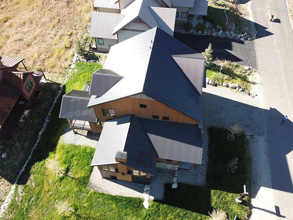 Aerial view of a modern wooden house with a dark metal roof, surrounded by green grass, neighboring houses, a gravel driveway, and a paved road.