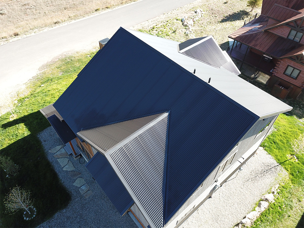 Aerial view of a house with a dark, corrugated metal roof featuring multiple peaks and slopes, surrounded by green grass, a gravel driveway, and a nearby road.