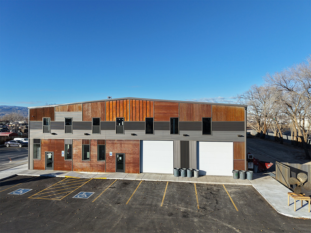 A modern industrial building with wood and metal siding features two large white garage doors, several windows, and a row of garbage bins. The parking lot in front has marked spaces, including handicapped spots.