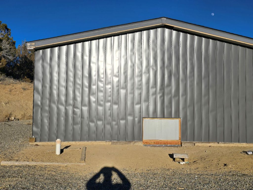 A corrugated metal building wall with a small framed opening near the ground, surrounded by dirt and gravel; a person’s shadow is visible in the foreground, and a clear blue sky with a faint moon is in the background.