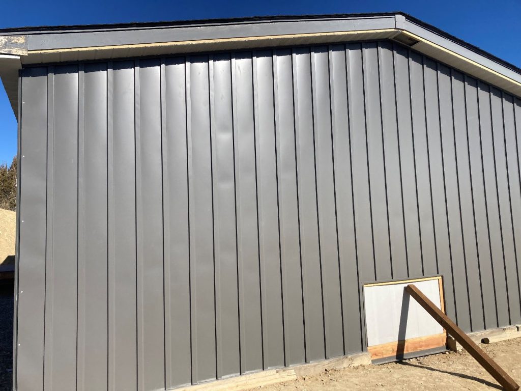 A gray metal-sided building with vertical panels, a white trim along the roof, and a small rectangular white and wood-framed section near the ground supported by a propped wooden beam. The sky is clear and blue.