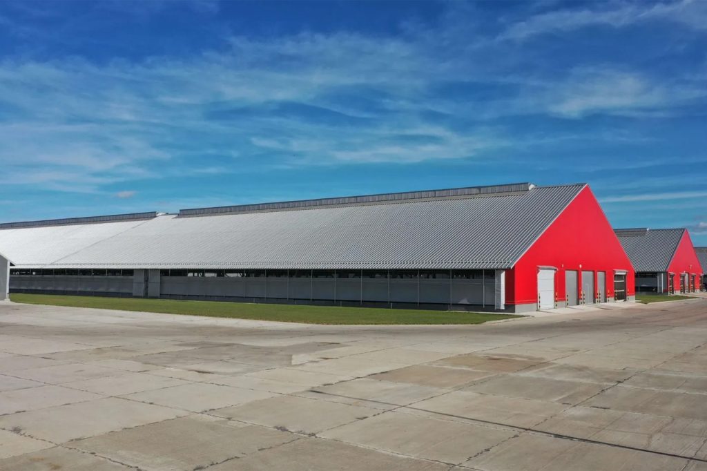 Large modern poultry farm building with bright red end walls, a gray slanted metal roof, and open sides, set on a wide concrete area under a blue sky with scattered clouds.