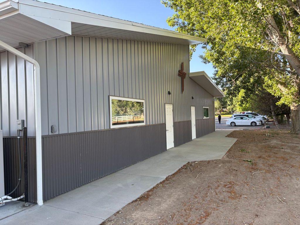 A metal building with a white door and a cross above it, situated next to a sidewalk and trees. There are parked cars and more trees visible in the background.