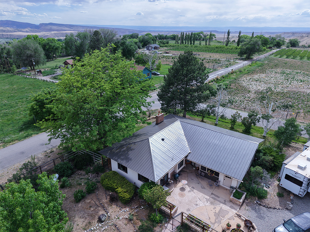 Aerial view of a house with a metal roof surrounded by trees and greenery, adjacent to a road, with farmland and fields stretching toward distant hills under a partly cloudy sky.