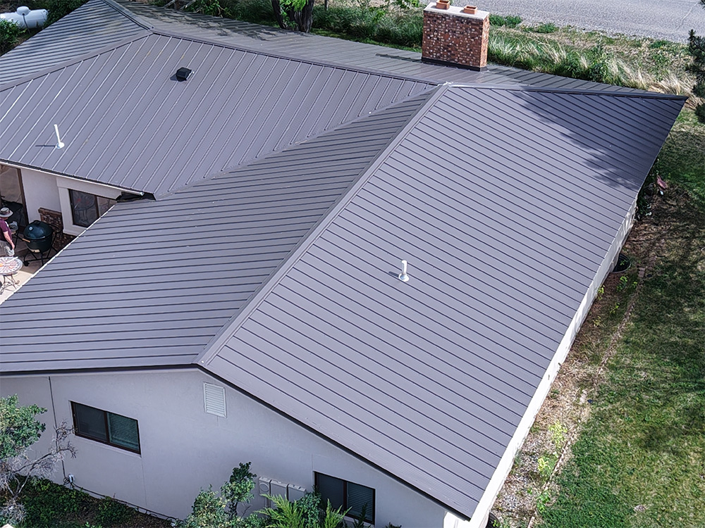 Aerial view of a house with a dark gray standing seam metal roof, surrounded by grass and some plants, with a brick chimney visible and part of a road in the background.