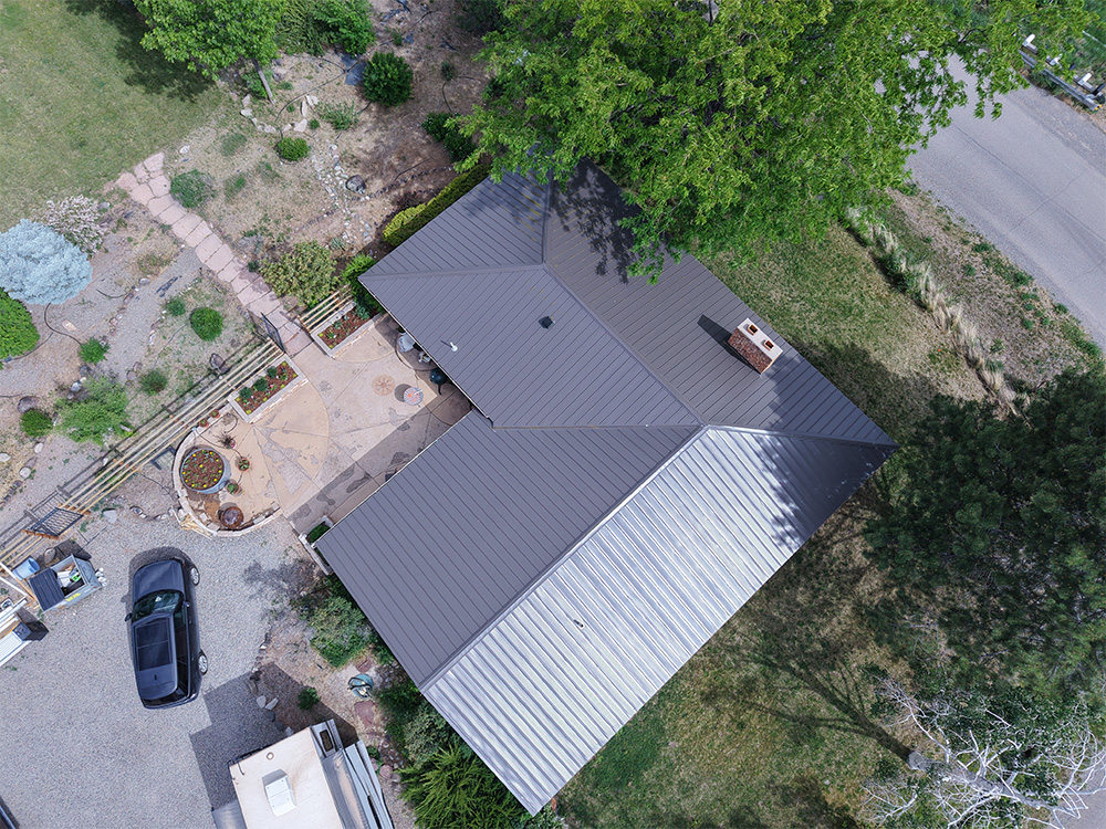 Aerial view of a house with a gray metal roof surrounded by trees, a garden, pathways, and a parked car in the driveway. The house sits near a road, and the yard includes landscaping features and plants.
