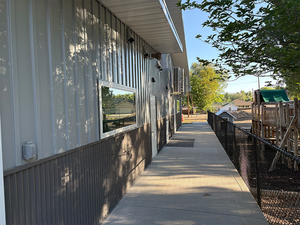 A concrete walkway runs alongside a metal building with a window. A black fence separates the path from a playground area with slides and climbing equipment. Trees cast shadows on the building and path.