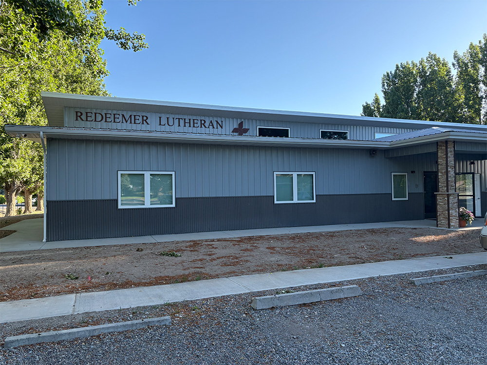 A gray building with a sign reading "Redeemer Lutheran" above the windows, featuring a cross symbol. There are trees nearby and a sidewalk with some gravel and dirt in front of the building.
