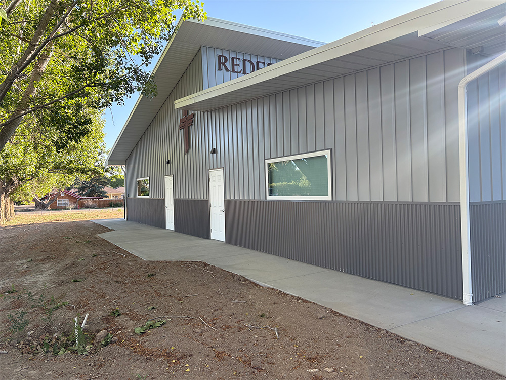 A gray metal building with a slanted roof, white doors, and a cross on the wall; a concrete walkway runs alongside the building, with dirt and sparse vegetation nearby.