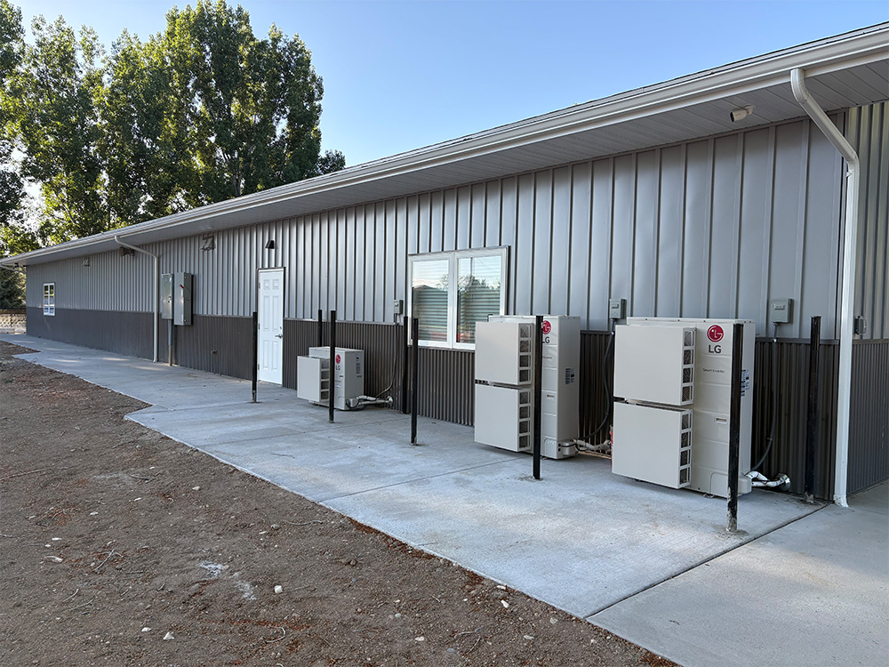 Exterior view of a long metal building with several LG HVAC units installed along the concrete walkway beside it, under a clear blue sky with trees in the background.