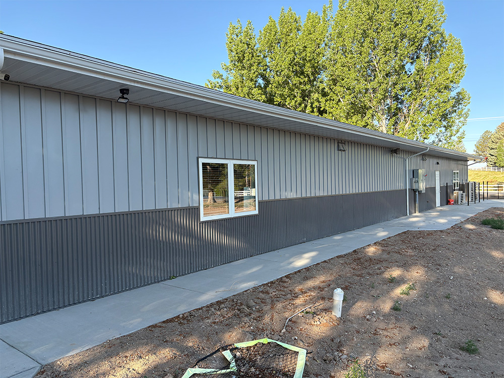 A long, metal-sided building with a gray exterior, large windows, and a concrete walkway alongside bare earth, with green trees and a clear blue sky in the background.