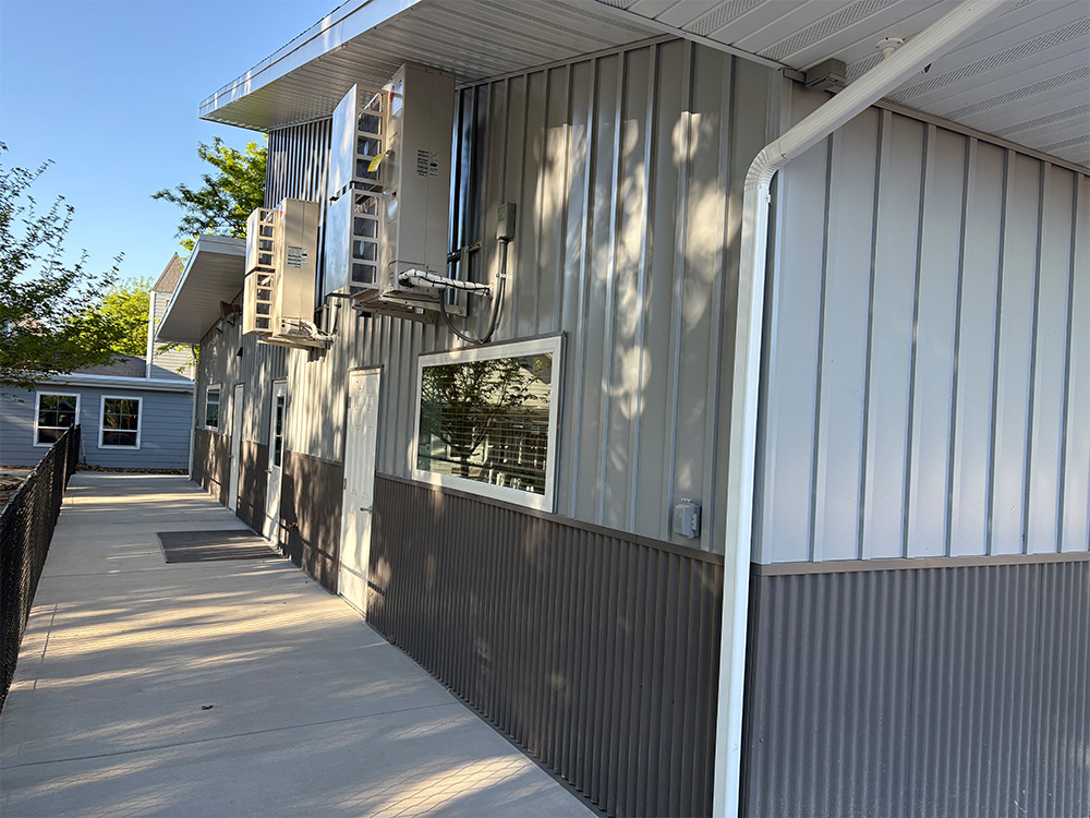 A long, metal-sided building with several air conditioning units mounted high on the wall, a window with reflections, and a sidewalk bordered by a black metal fence on a sunny day.