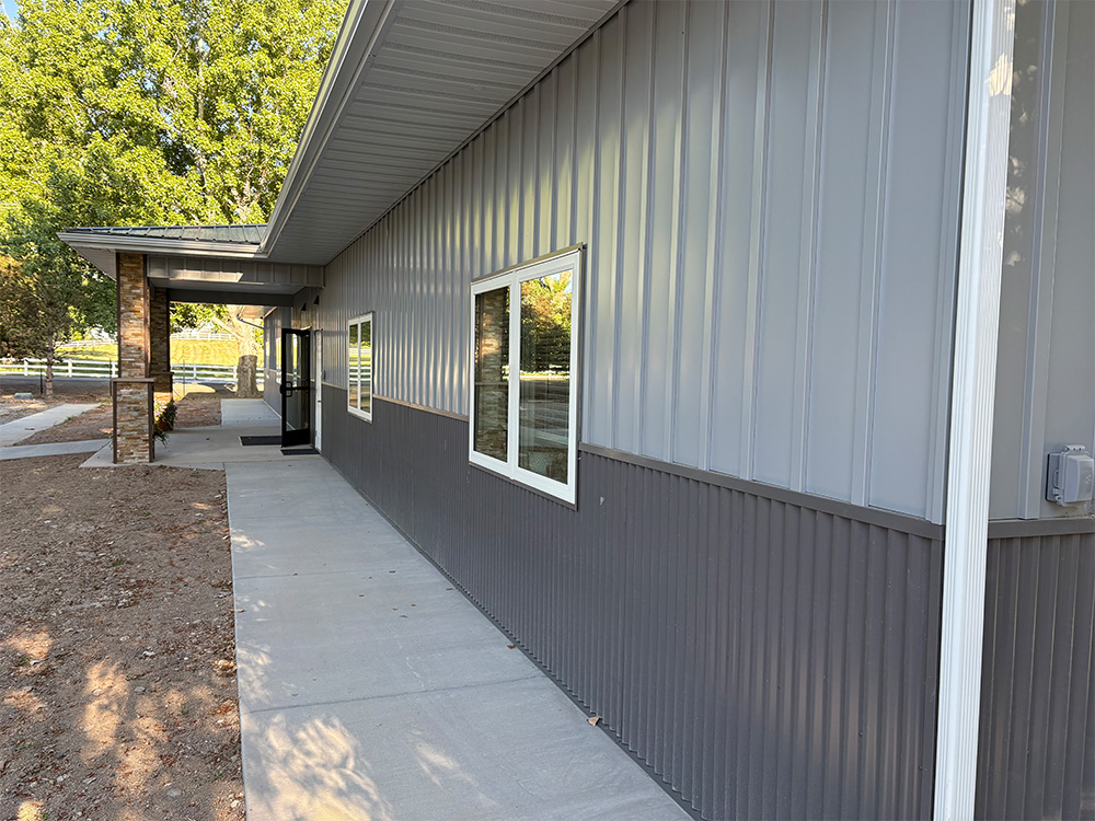A modern building with gray metal siding, large windows, and a covered entrance. The concrete walkway runs alongside the building, and trees and a fence are visible in the background.