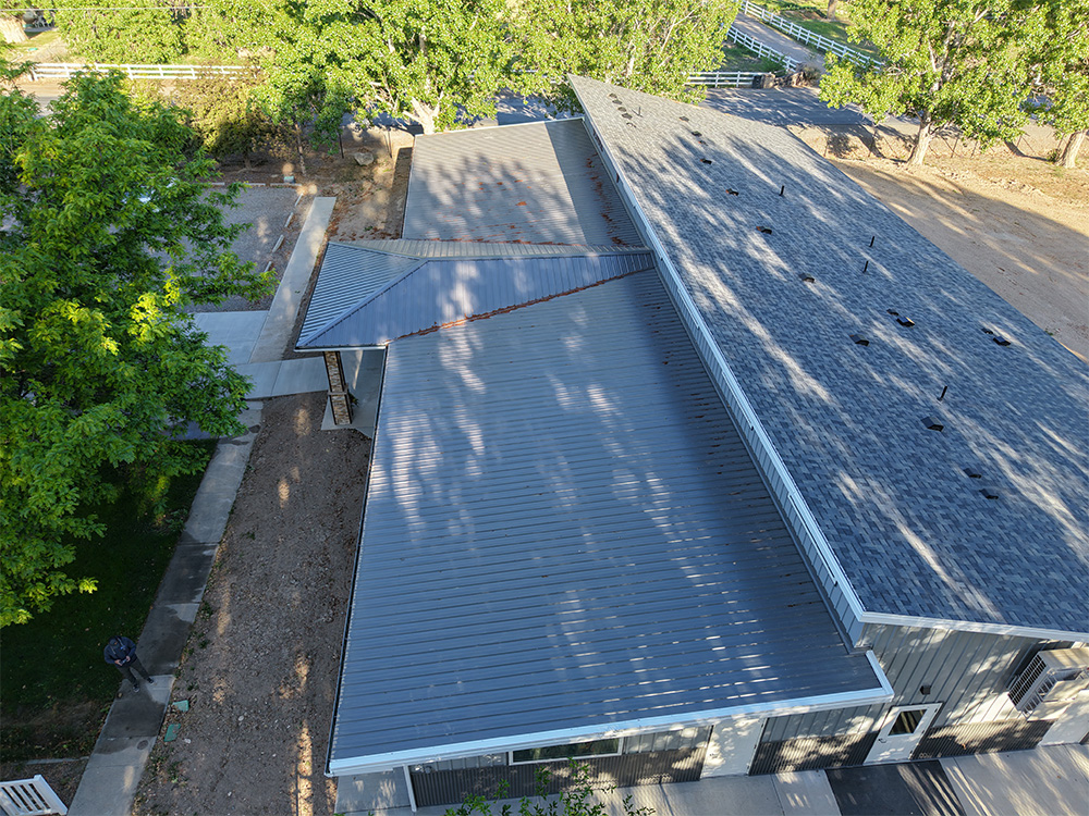 Aerial view of a building with a large metal roof, surrounded by trees and a dirt area, with a paved walkway leading to the entrance.