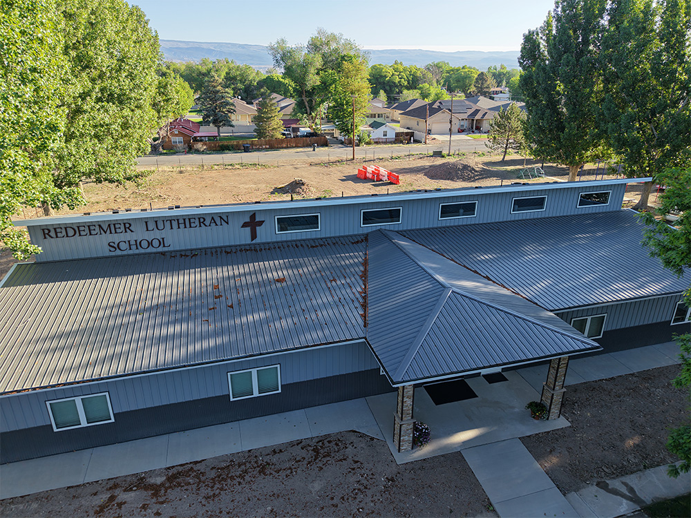 Aerial view of Redeemer Lutheran School building with a gray metal roof, surrounded by trees and a residential neighborhood in the background.