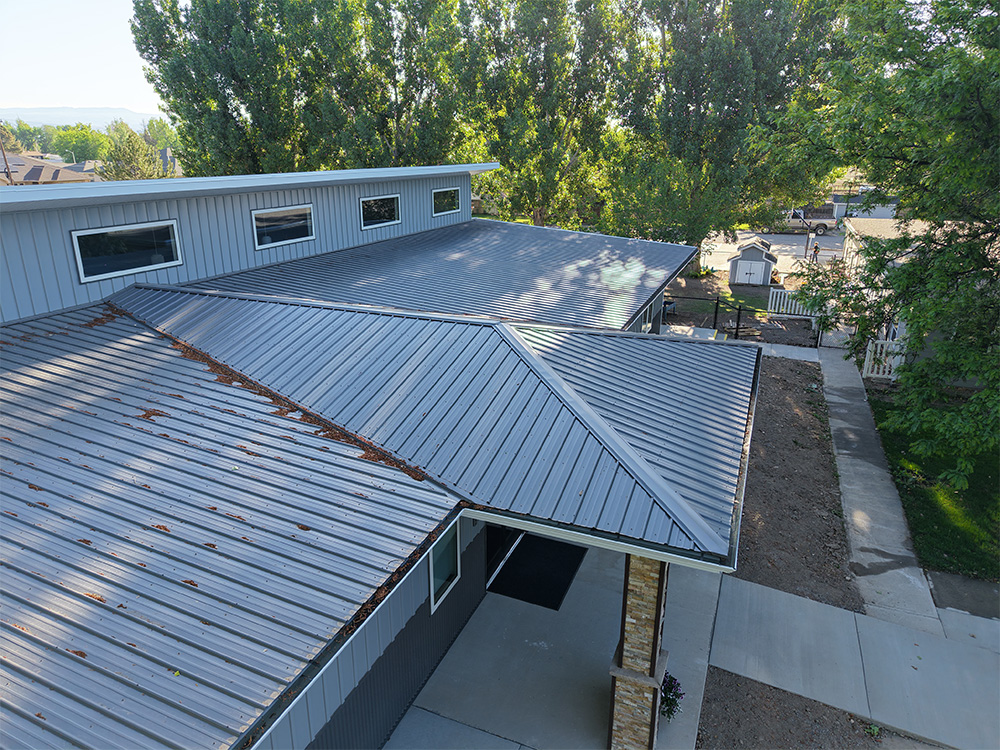 Aerial view of a modern building with a gray metal roof, surrounded by trees and concrete walkways, with sunlight filtering through the leaves.